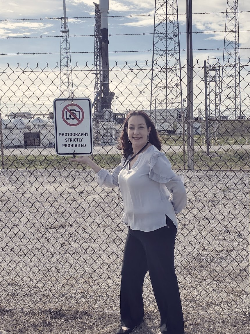 Meagan Crawford resting a hand on a sign reading Photography Strictly Prohibited, with a rocket on the pad behind the fence.